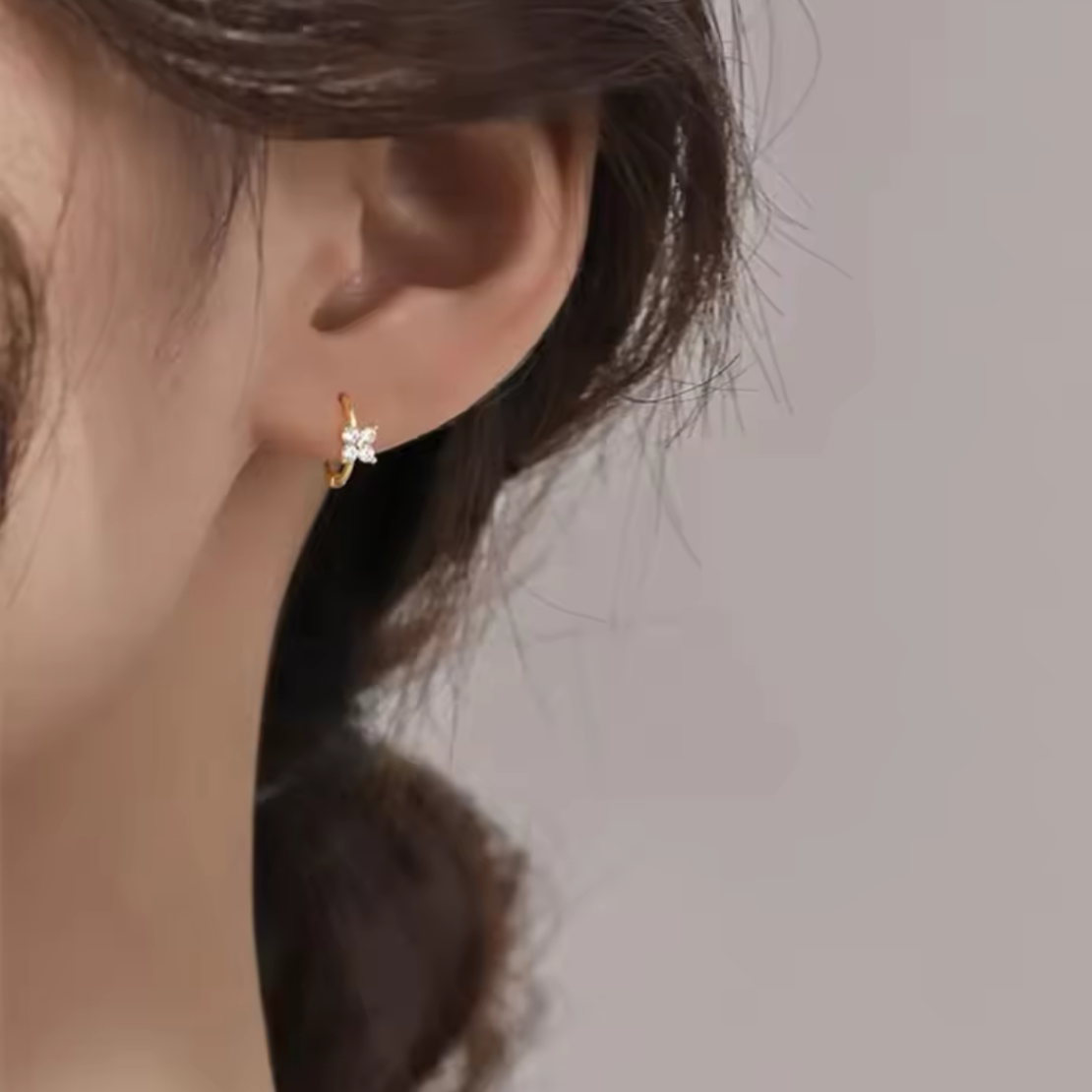 Close-up of an ear wearing a gold earring with a clear gemstone on a neutral background